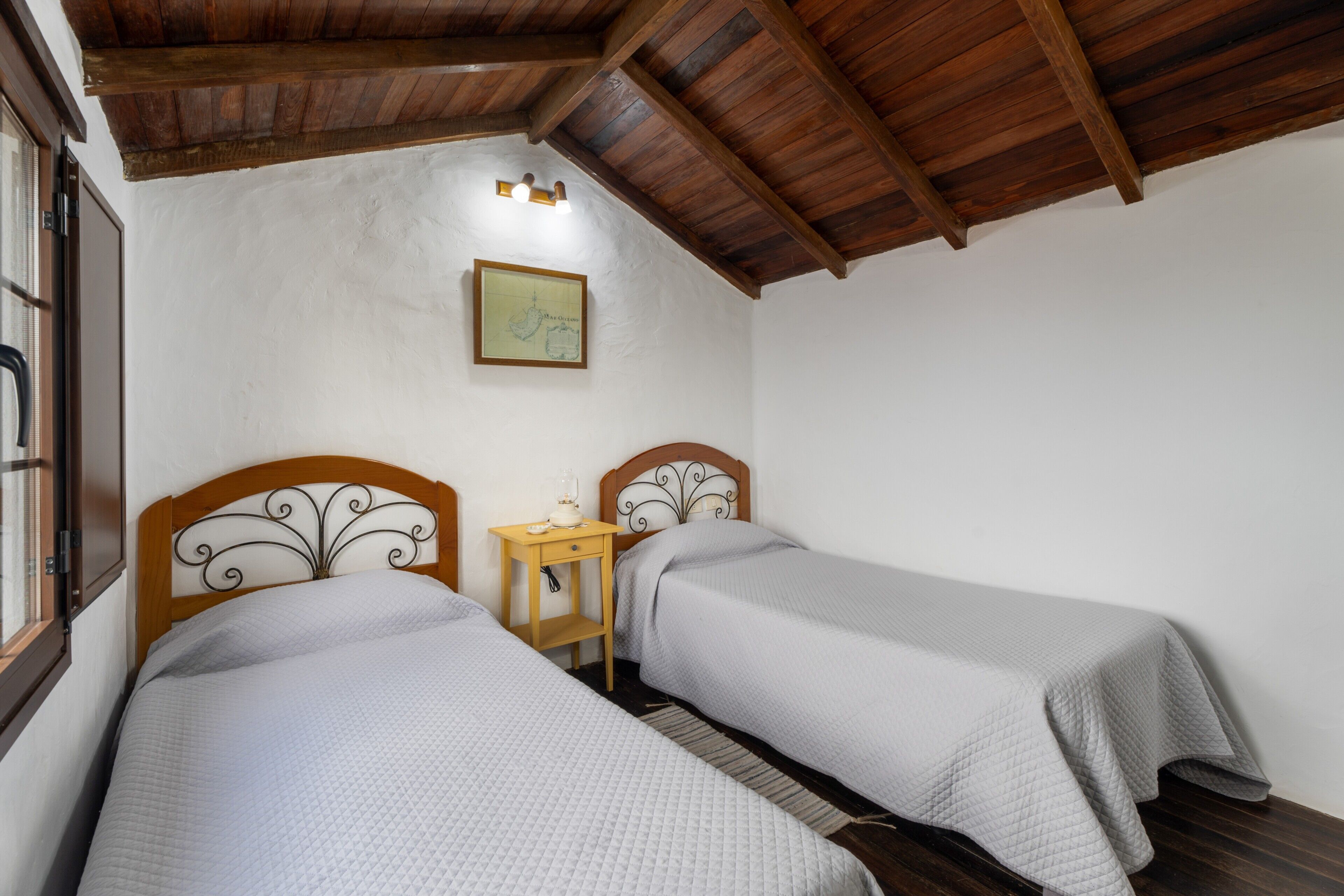 A room with two beds, wooden headboards, a small bedside table, a framed map on the wall, and a window.