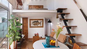 Un interior moderno con una mesa de comedor, sillas, una planta en maceta, un cuadro de peces, una escalera y vistas a través de una ventana.