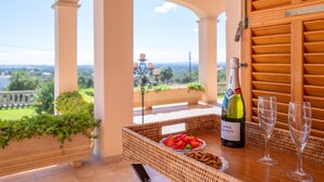 A wicker table with a bottle of wine, two glasses, and a bowl of fruit on a balcony overlooking a scenic landscape.