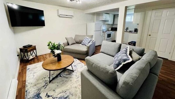 Ein Wohnzimmer mit einer Couch, einem Couchtisch und einem Flachbildfernseher. Im Hintergrund ist eine Küche zu sehen.