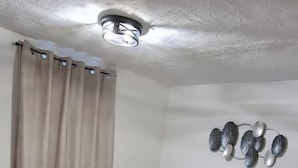 Ein Schlafzimmer mit einem Bett, einem Kopfteil, einer Deckenleuchte, einer Wanddekoration und einem Fenster mit Vorhängen.