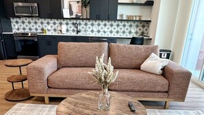 A modern living room with a brown sofa, a round coffee table with a vase, and a kitchen in the background with dark cabinets and a tiled backsplash. - “Freiheit Loft” Lush Couple’s Getaway by Gruene (New Braunfels)