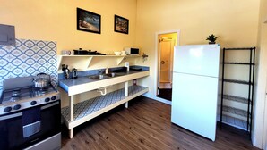 A compact kitchen with a stove, microwave, and refrigerator. There are two framed pictures on the wall, a small shelf with kitchenware, and a door leading to a closet. - Sunny 1 bedroom in Esperanza close to everything (Vieques)