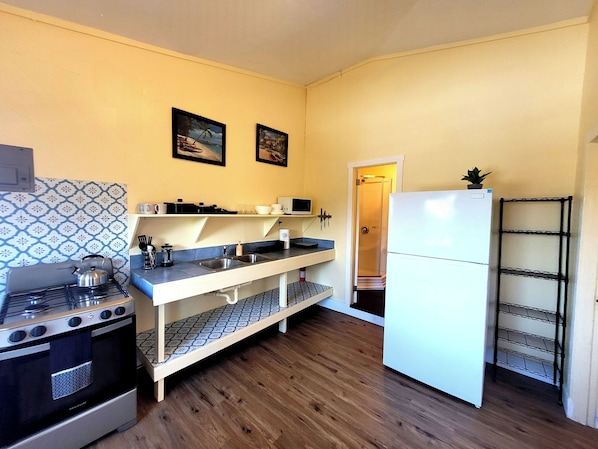 Una cocina compacta con estufa, microondas y refrigerador. Hay dos cuadros enmarcados en la pared, un estante pequeño con utensilios de cocina y una puerta que da a un armario.