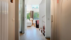 En hall som leder till ett sovrum med en säng, en stol och ett bord. Det finns ett fönster med gardiner och utsikt över grönområden utanför.