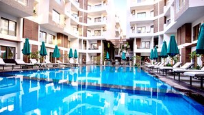 Et moderne hotel med en central pool, flere balkoner og udendørs siddeområder med parasoller.