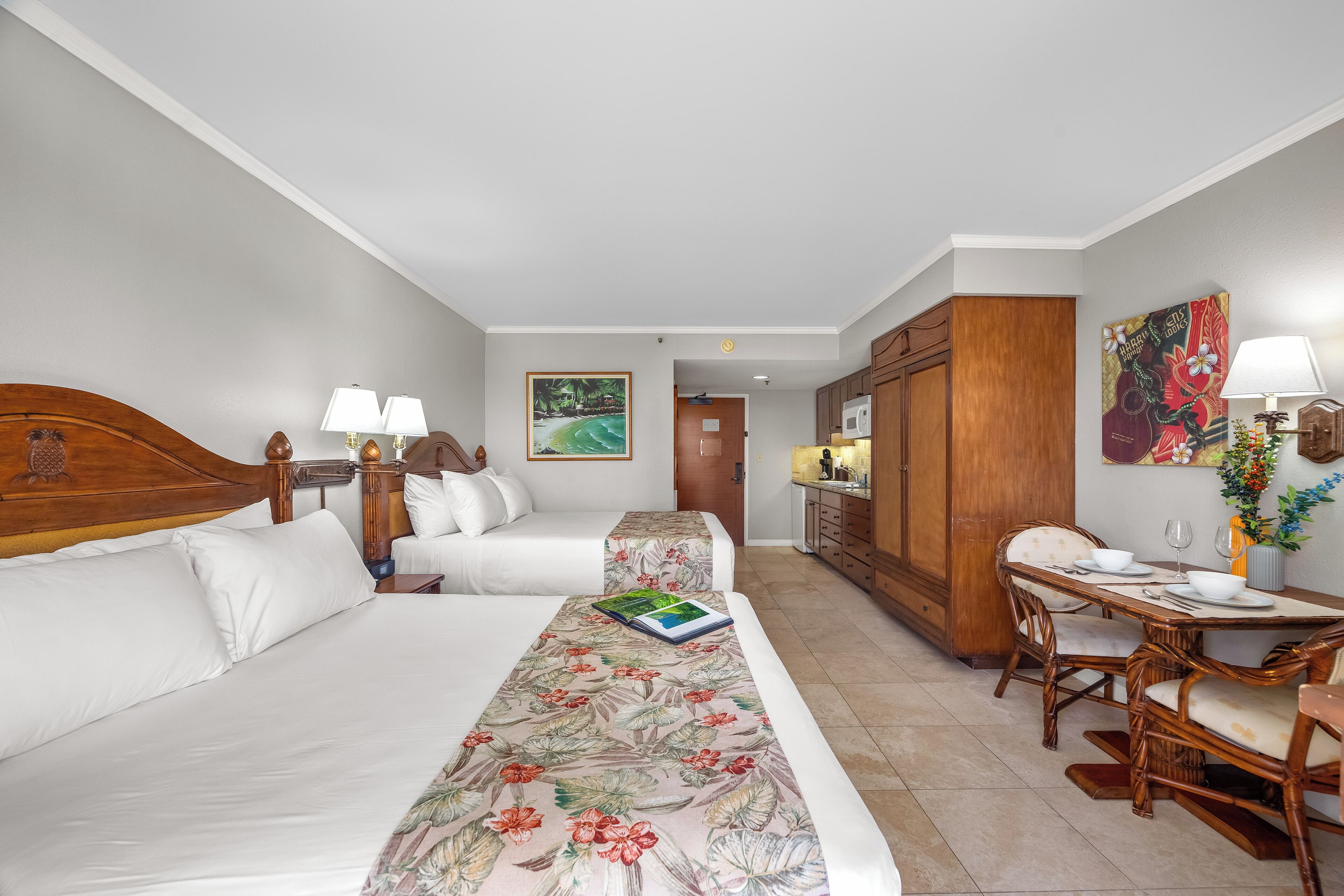 A hotel room with two beds, a dining table with chairs, a kitchen area, and a painting on the wall.