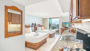 A hotel room with two beds, a kitchen area with a sink and a coffee maker, and a large window offering a city view.