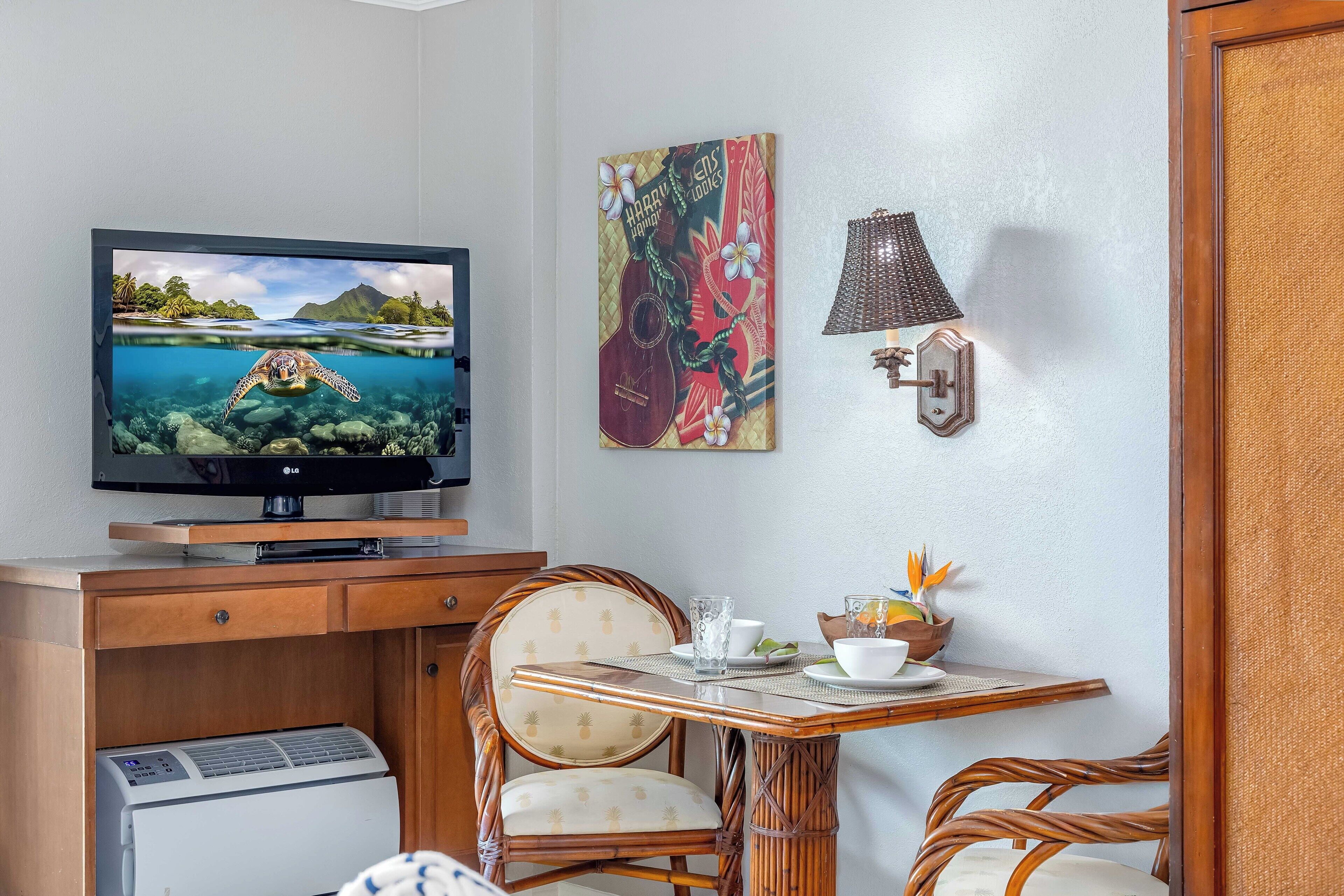 Habitación con televisión mostrando una escena tropical, una mesa de comedor con sillas, un refrigerador pequeño y una lámpara fijada a la pared.