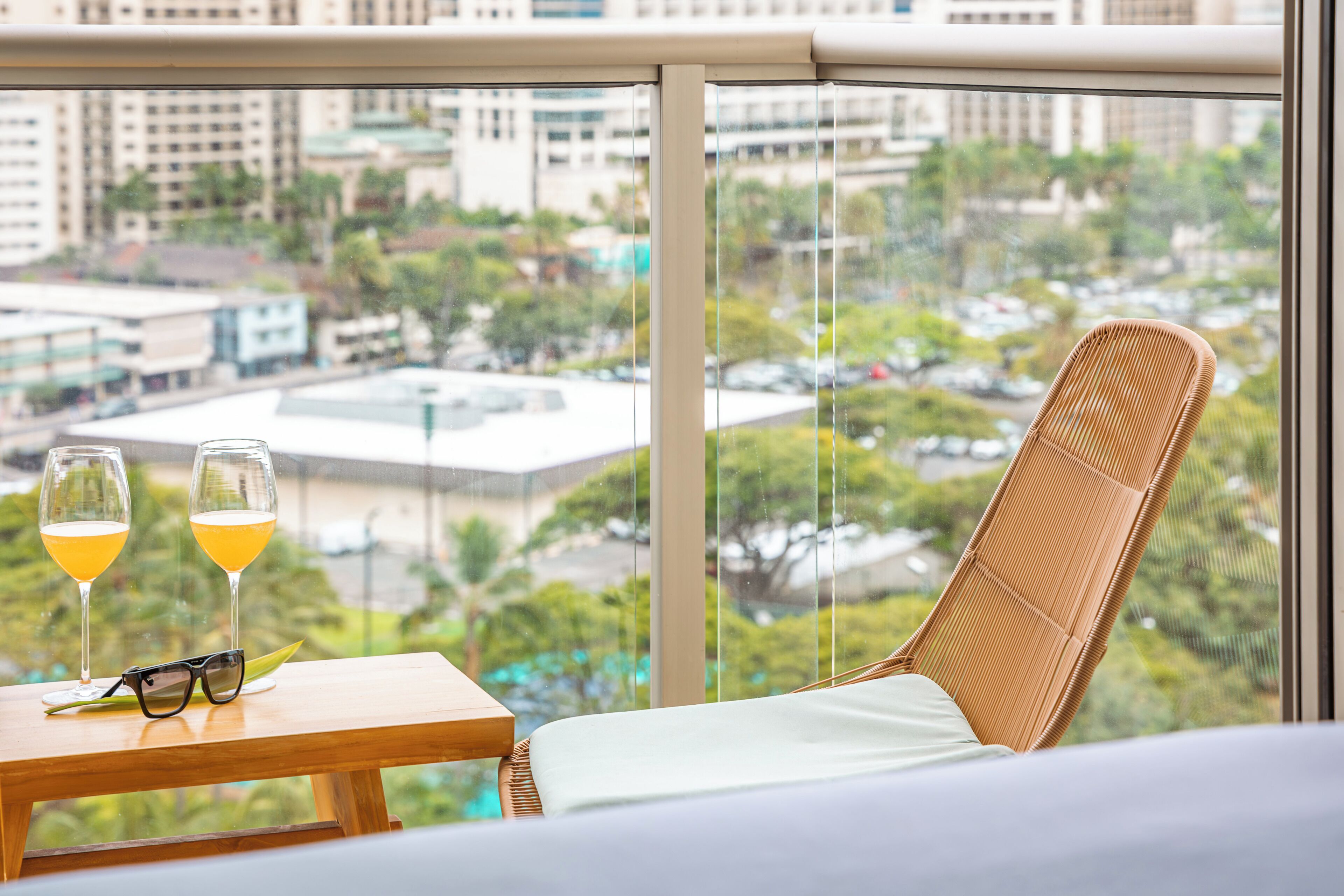 Un balcón con vista a un paisaje urbano, una silla de mimbre, una mesa con dos vasos de jugo de naranja y un par de gafas de sol.