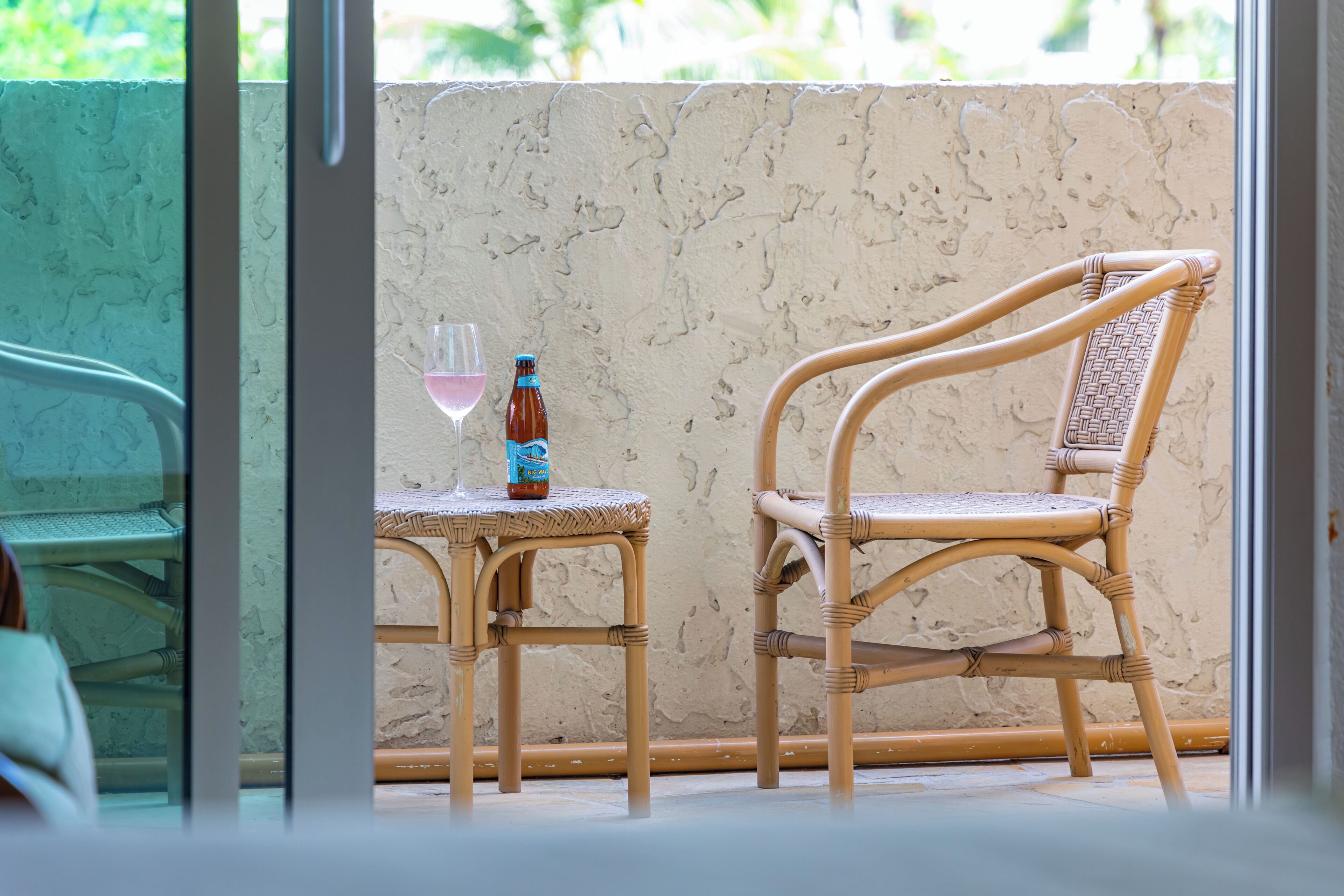 A wicker chair and table with a bottle and glass on a balcony with a textured wall and a view of greenery.
