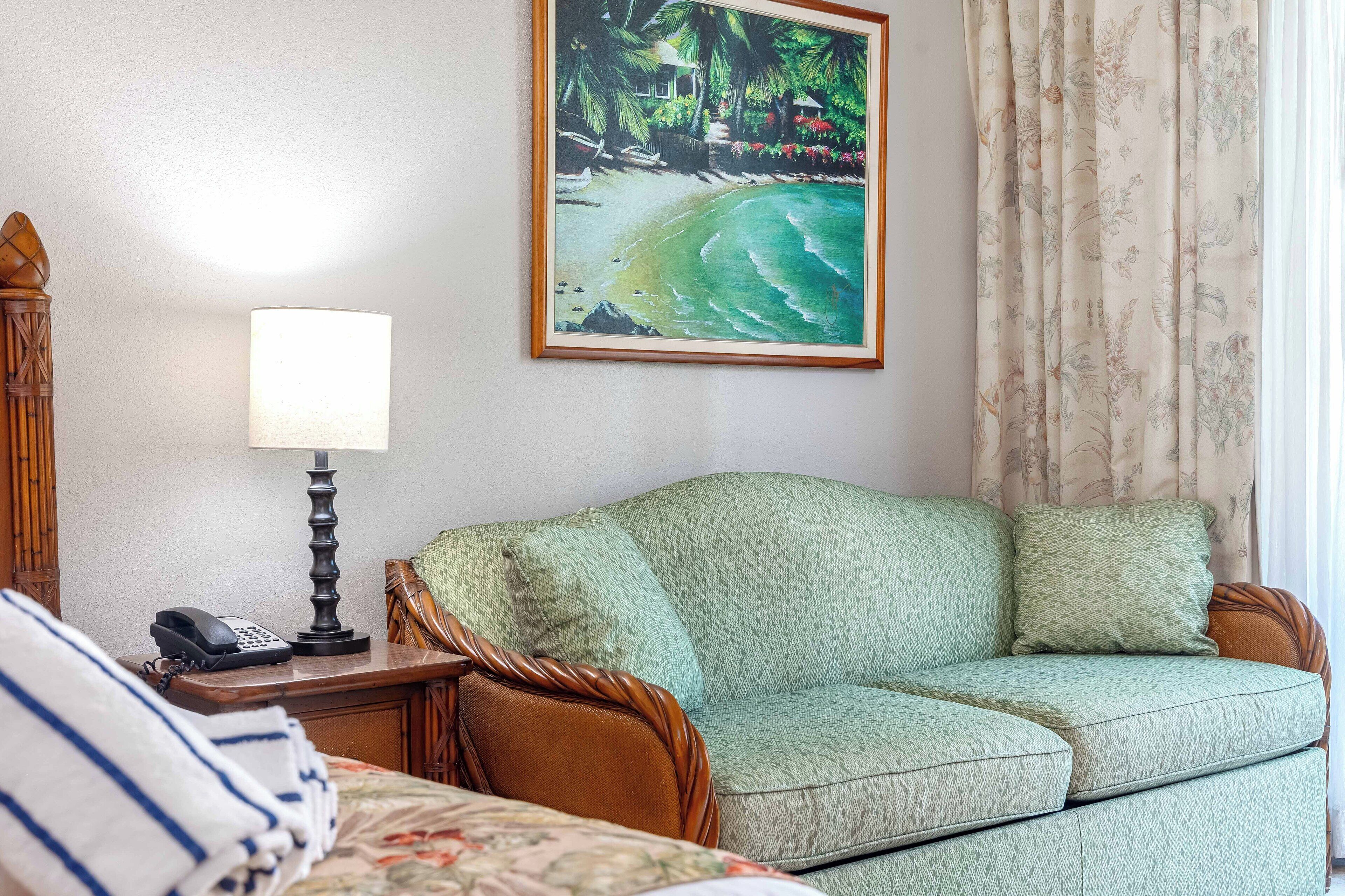 A room with a green sofa, a wooden nightstand with a lamp, a framed painting of a beach scene, and a bed with striped bedding.