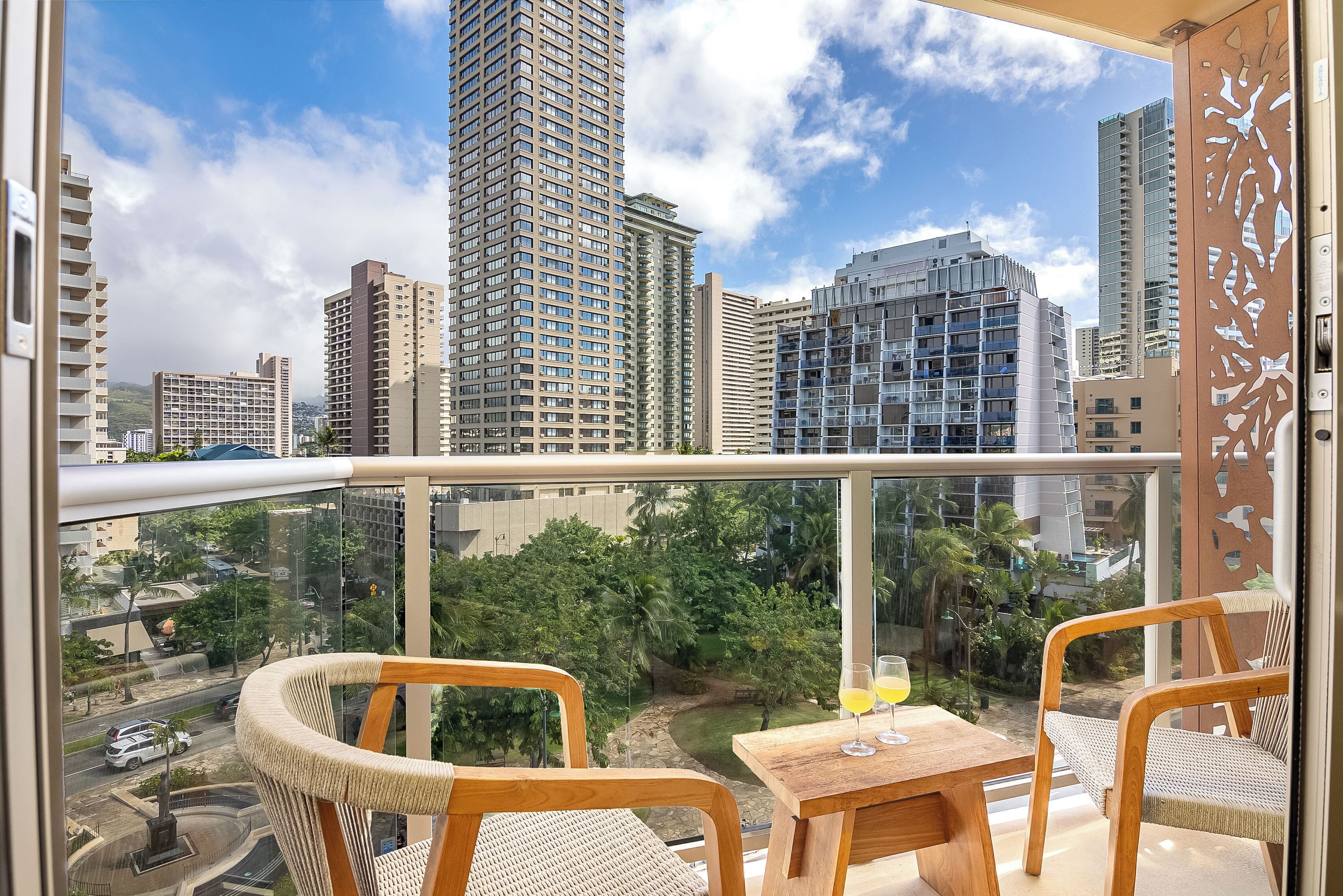 A balcony with two wicker chairs and a small table, overlooking a cityscape with tall buildings and greenery.