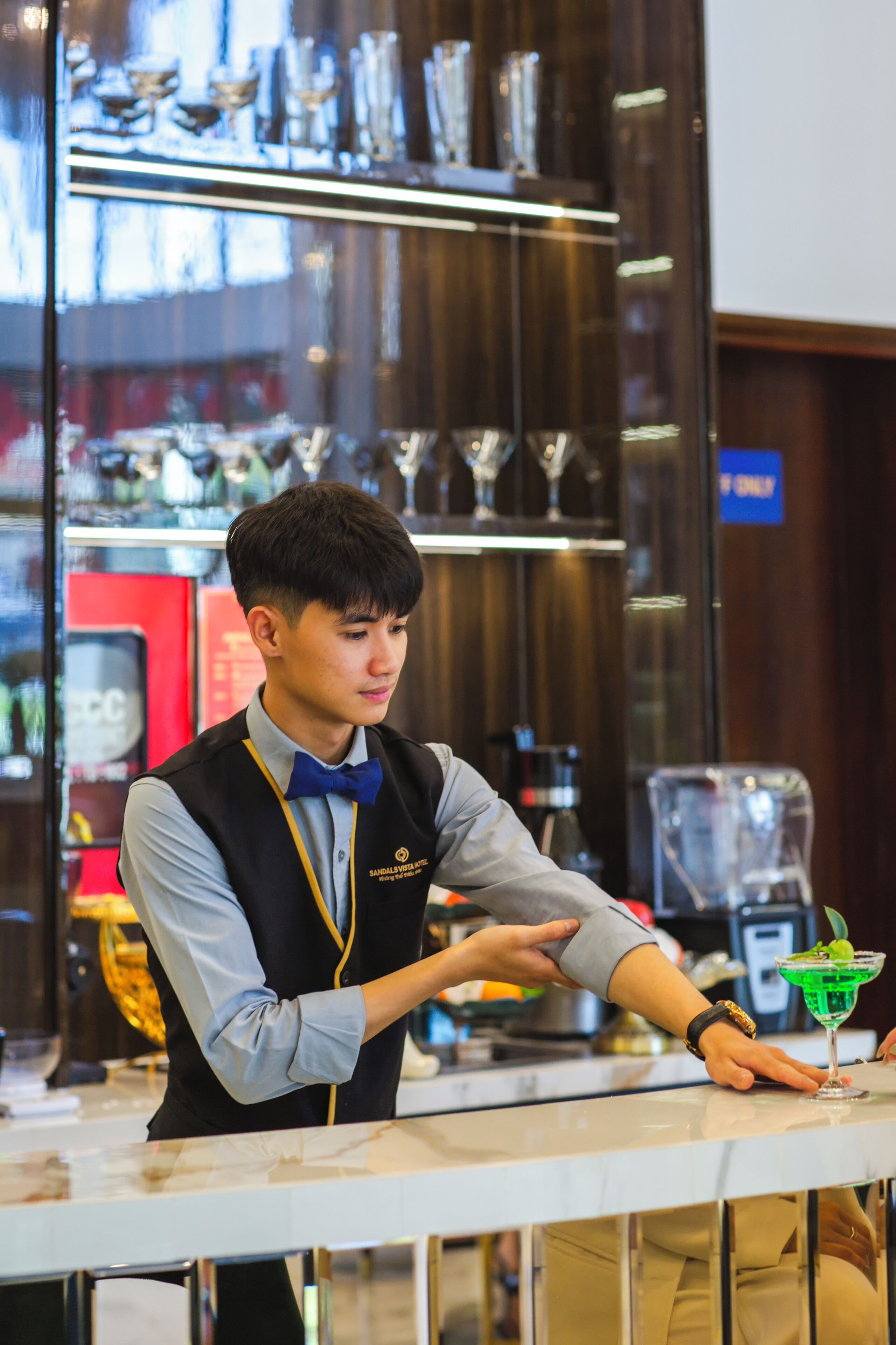 A bar area with a bartender, a glass of green cocktail, and shelves with glasses and bottles.