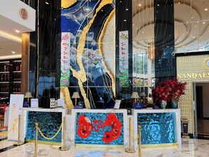 Eine luxuriöse Lobby mit einer großen, reflektierenden Wand, die eine Frau in einem Kleid zeigt, einem Empfangsbereich und dem Eingang eines 'SANDALS' Geschäfts.