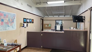 A reception area with a counter, a map on the wall, a telephone, and a television mounted on the ceiling. - American Inn Motel (Pratt)