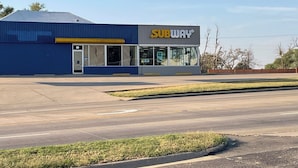 A Subway restaurant with a blue exterior and a large parking lot in front. - American Inn Motel (Pratt)