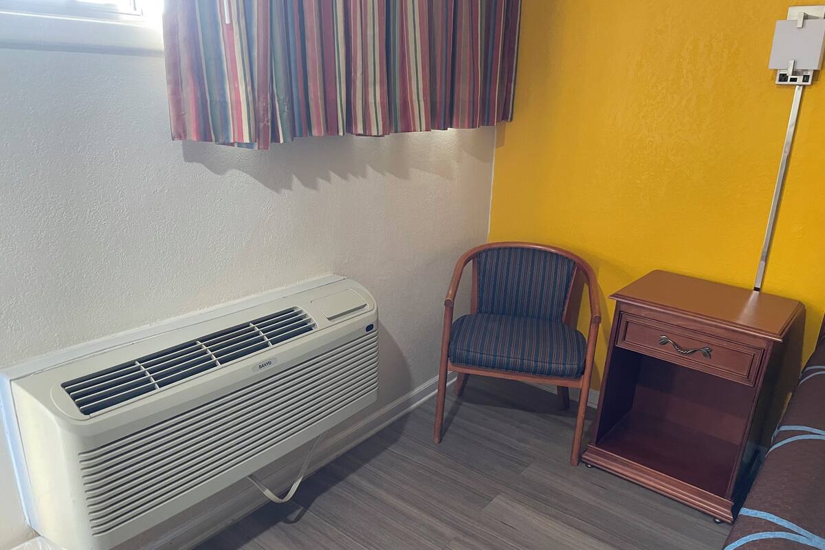 A room with a white air conditioner, a striped curtain, a wooden nightstand, and a chair.