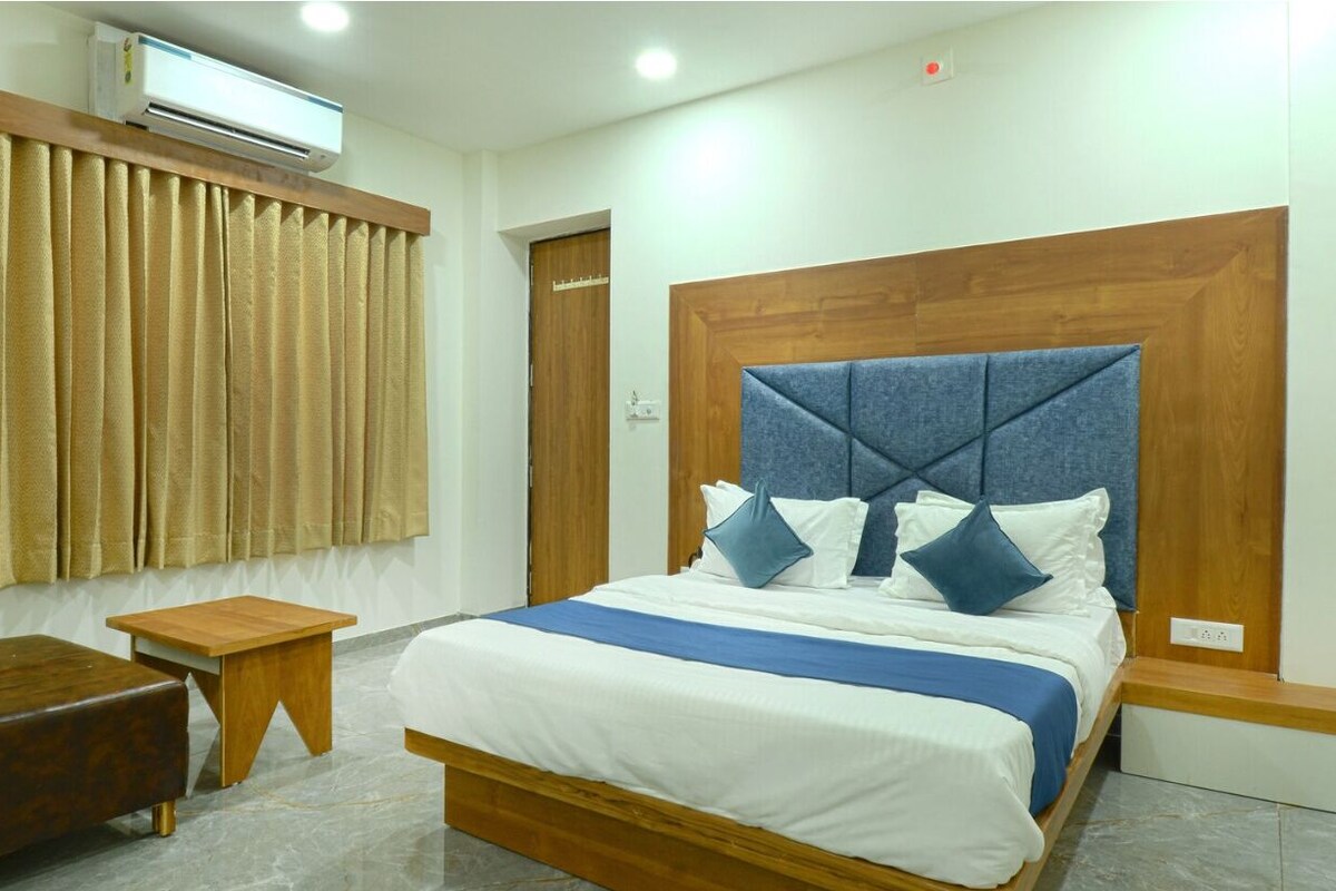 A hotel room with a bed, a wooden bench, a small table, and a window with curtains.