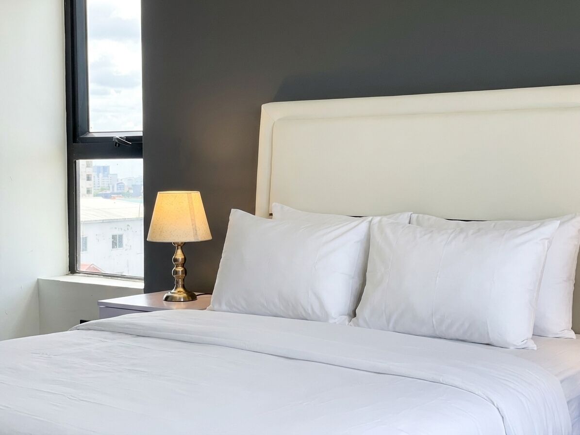 A neatly made bed with white linens, a bedside lamp, and a window with a view of buildings.