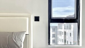 A bedroom with a bed, a nightstand, a window with a view of buildings, and a thermostat on the wall.