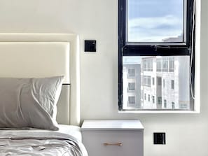 Ein Schlafzimmer mit Bett, Nachttisch, Fenster mit Blick auf Gebäude und einem Wandthermostat.