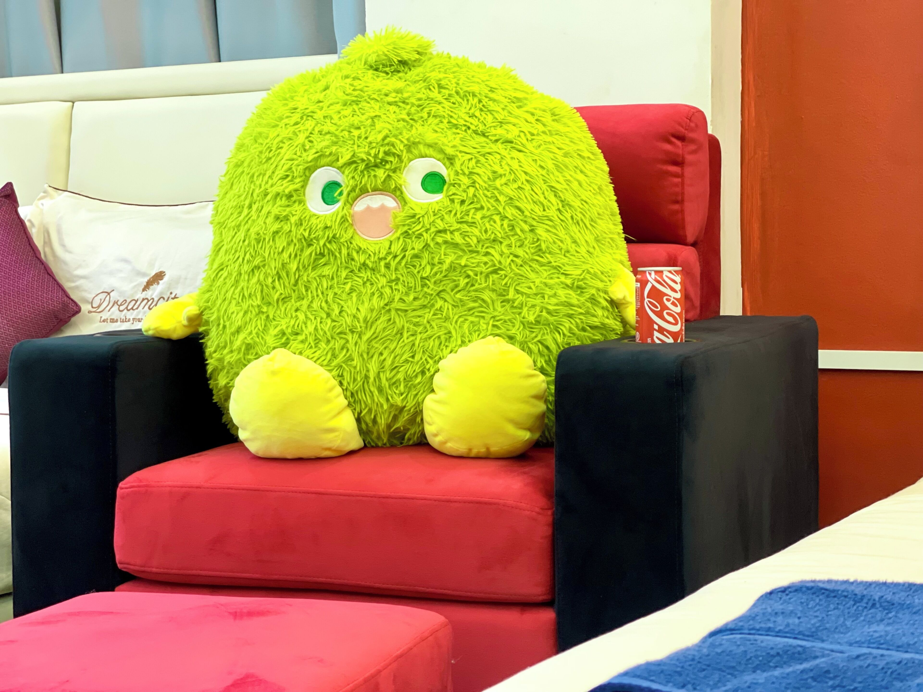 A plush toy with a green body and yellow feet seated on a red cushioned chair.