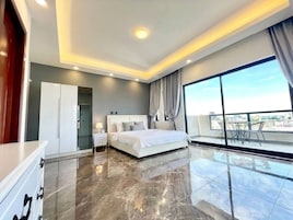 Quarto moderno com uma cama grande, um guarda-roupa, uma mesa de cabeceira e vista da cidade através de uma janela ampla.