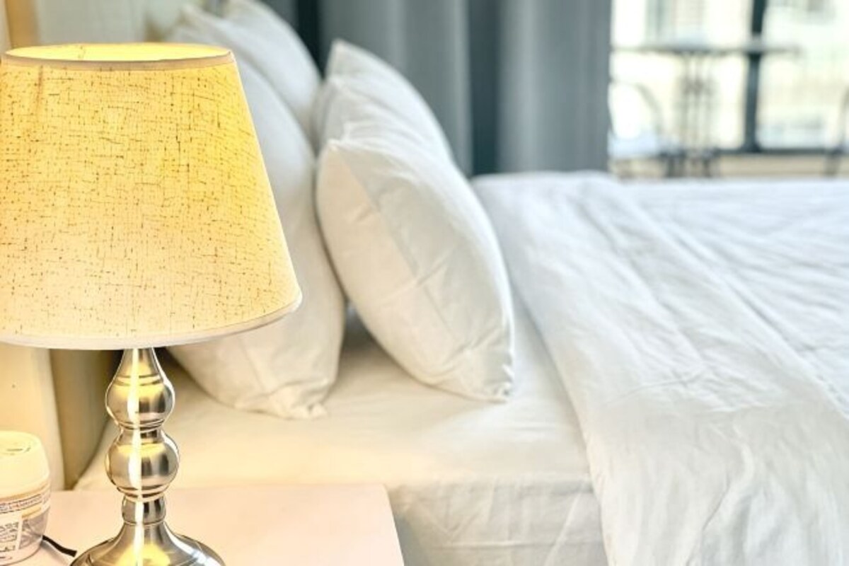 A bed with white bedding, a lamp on a bedside table, and a window in the background.