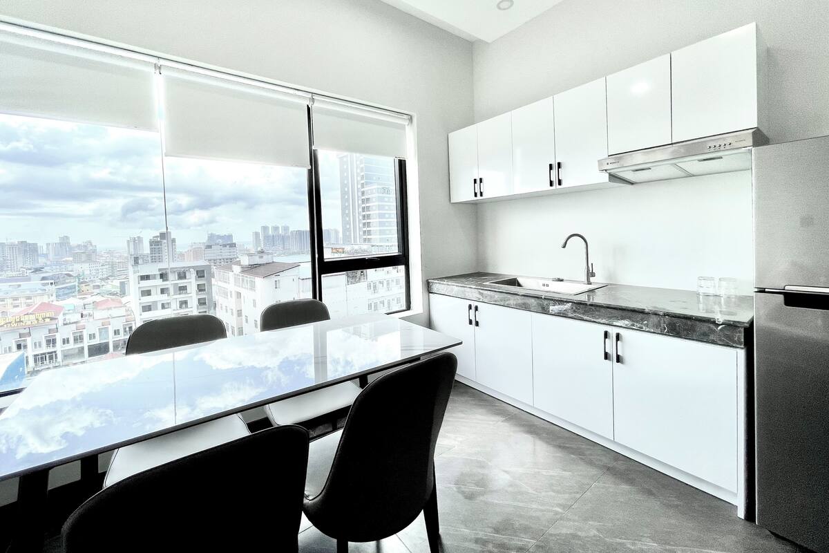 Una cocina moderna con un ventanal grande, armarios blancos, un refrigerador de acero inoxidable y un comedor con mesa y sillas de vidrio.