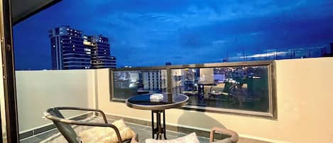 Un balcón con dos sillas de mimbre y una mesita, con vistas a un paisaje urbano al atardecer.