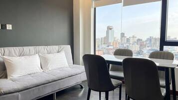 A modern living room with a grey sofa, two black chairs, and a dining table. A large window offers a city view.