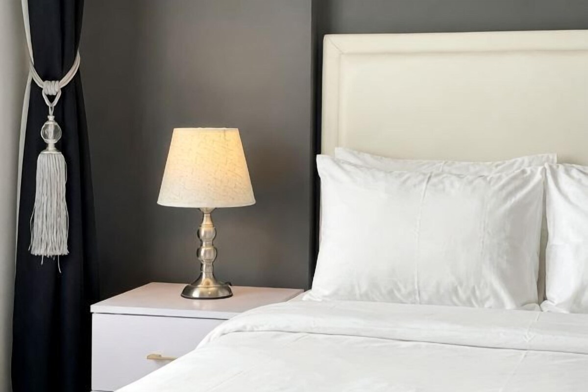 A neatly made bed with white linens, a bedside lamp with a white shade, and a dark wall in the background.