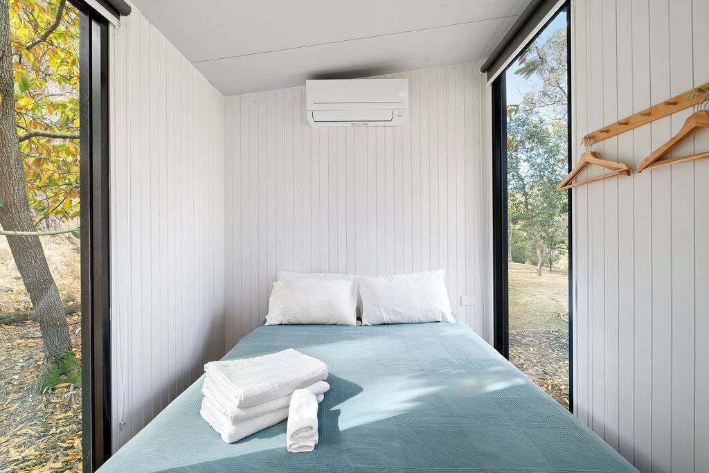A small, single-bed room with a window, a wall-mounted air conditioner, and a stack of folded towels on the bed.