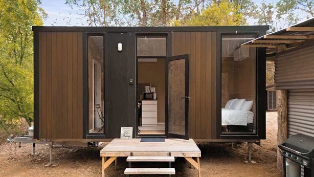 Una cabaña compacta con fachada de madera, un interior visible con cama y una pequeña kitchenette, y un juego de escalones exteriores que conducen a la entrada.