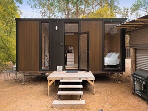 Une petite cabane au revêtement extérieur en bois, avec un intérieur visible comprenant un lit et une petite kitchenette, ainsi qu’un ensemble de marches extérieures menant à l’entrée.
