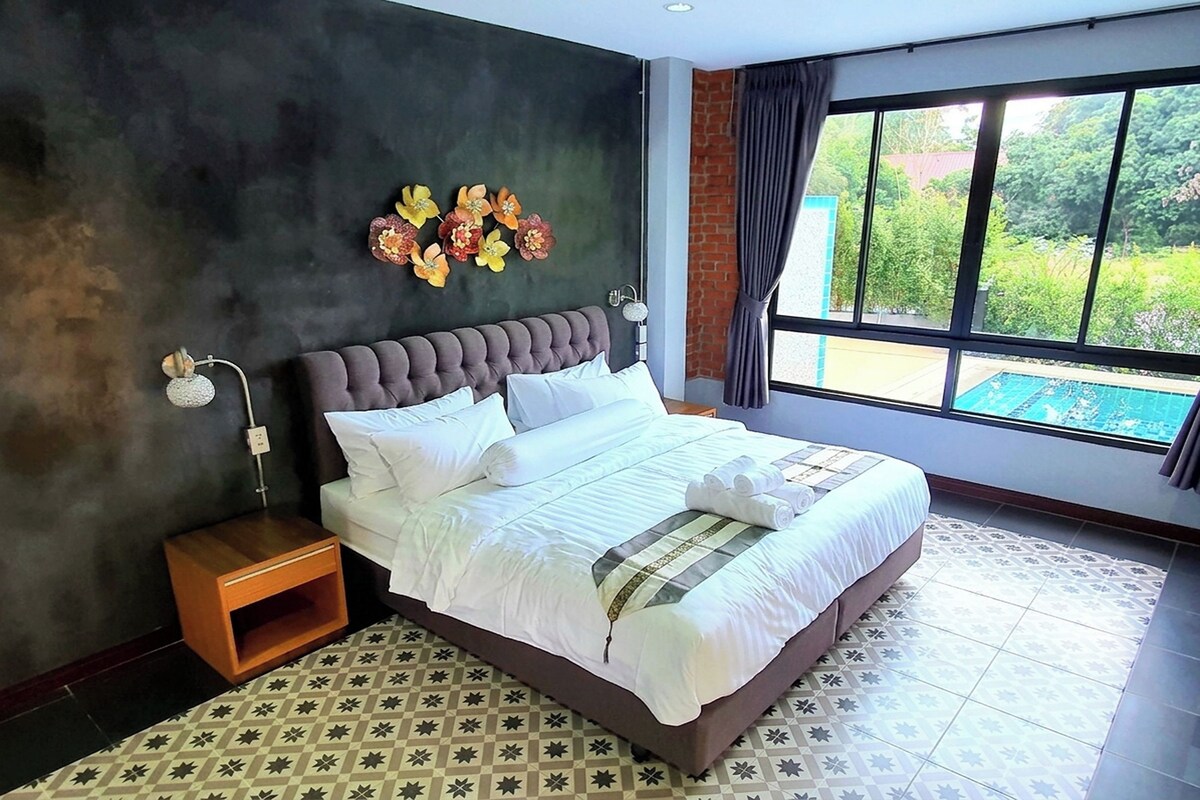 A hotel room with a large bed, a nightstand, a wall-mounted lamp, and a view of a swimming pool through a window.