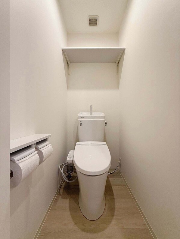 A small, white bathroom with a toilet, a toilet paper holder, and a shelf above the toilet.