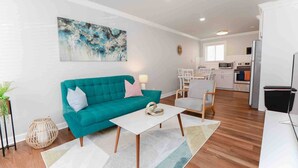 A modern living room with a teal sofa, a white coffee table, and a kitchen in the background.
