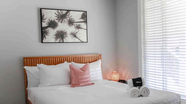 A neatly made bed with white and pink pillows, a framed artwork above it, and a bedside lamp.