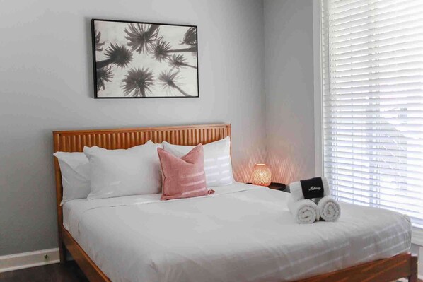 Una cama bien hecha con almohadas blancas y rosadas, una obra enmarcada encima y una lámpara de noche.