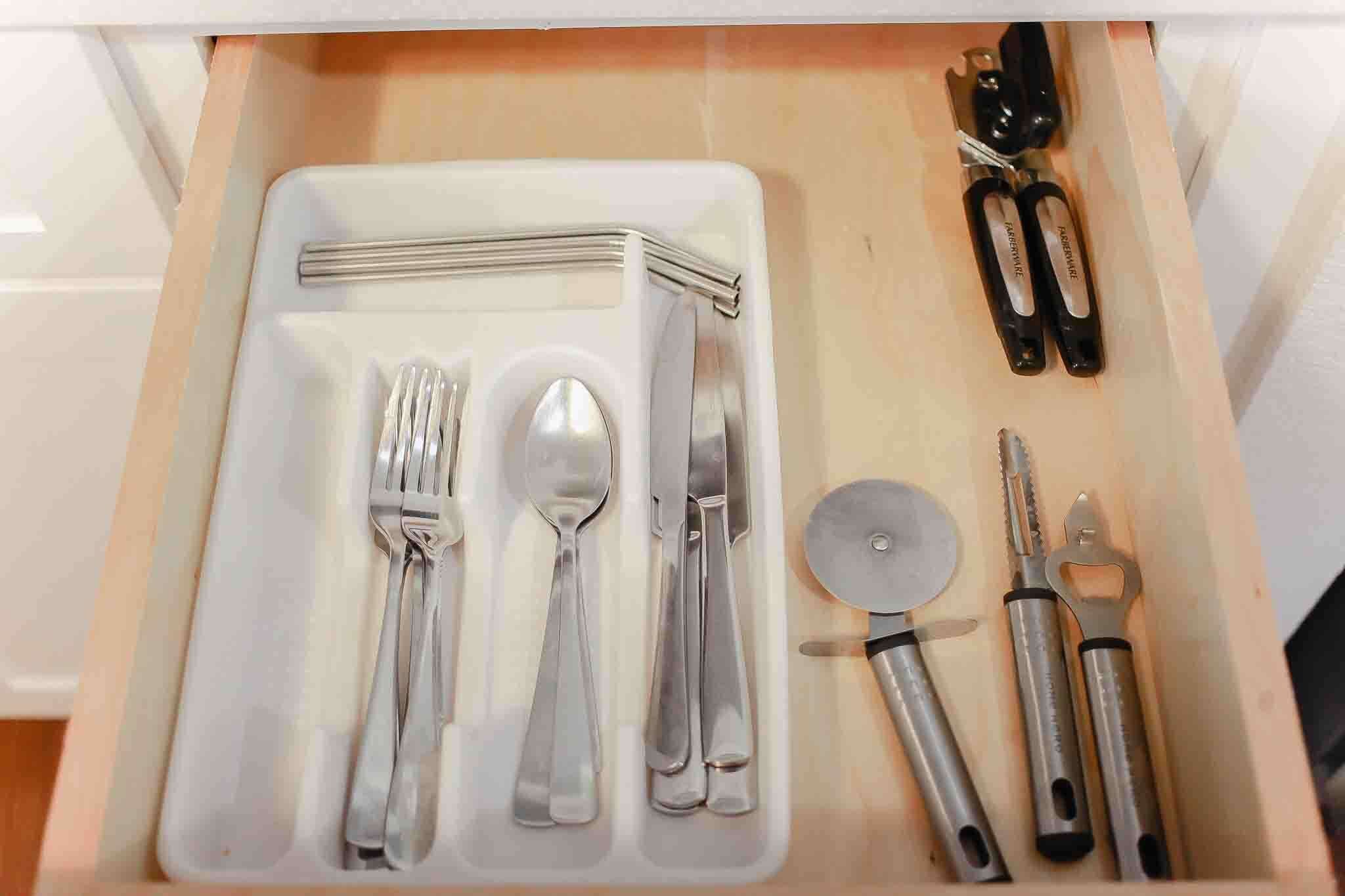A drawer containing a set of silverware, a spoon, a knife, a pizza cutter, and two black-handled tools.