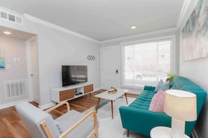 Un salon moderne avec un canapé de couleur turquoise, un téléviseur posé sur un meuble blanc, une petite table blanche et une lampe.