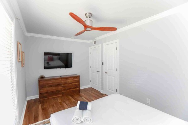 A bedroom with a wooden dresser, a TV mounted on the wall, a ceiling fan, and a bed with rolled towels.
