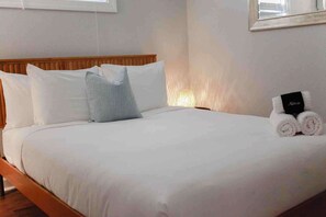 Una cama bien hecha con sábanas blancas y un cabecero de madera. Junto a la cama, una mesita con un cartel negro y toallas enrolladas.