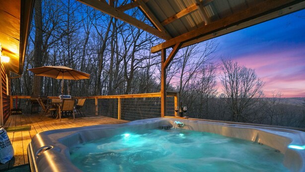 Un bain à remous avec une terrasse en bois et des chaises, entouré d’arbres et offrant une vue sur le coucher de soleil.