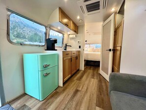 A compact kitchenette with a window, a small fridge, wooden cabinets, and a bed in the adjacent room.