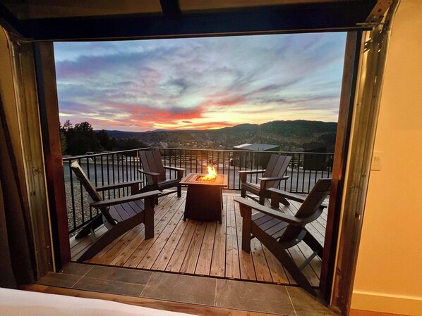 A deck with a fire pit and chairs, overlooking a scenic landscape with mountains and a sunset.