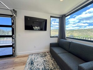 A room with a gray sofa, a mounted TV, a large window with a view of a mountainous landscape, and a wooden floor.