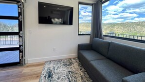 Ein Zimmer mit grauem Sofa, einem Wand-Fernseher, einem großen Fenster mit Blick auf eine Berglandschaft und Holzboden.